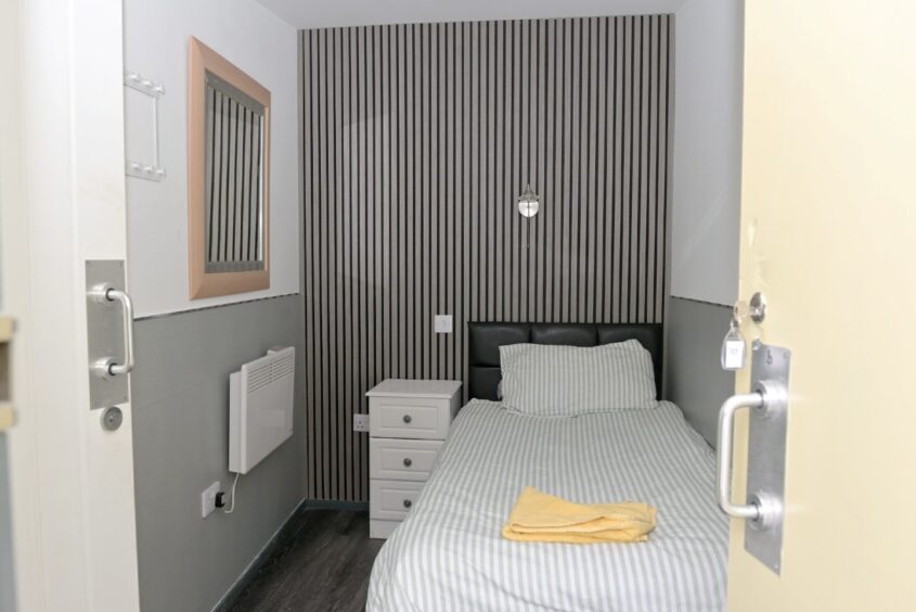 Image shows one of the single rooms at the hostel. The room is small. It has a single bed, a small set of drawers and a radiator. The decoration is grey wooden cladding.