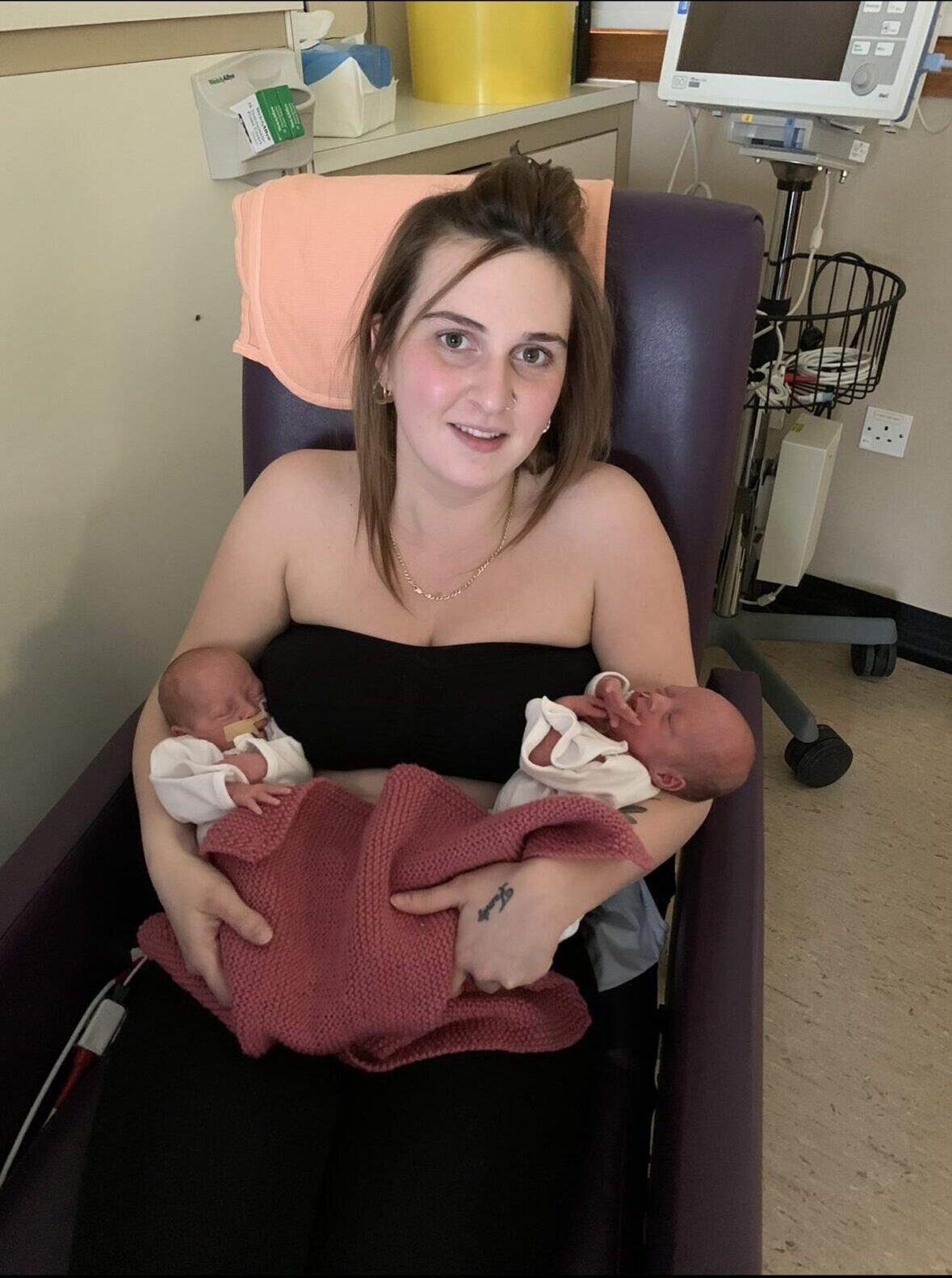 Tiny twins wrapped in blankets, being held one on each arm by their mum