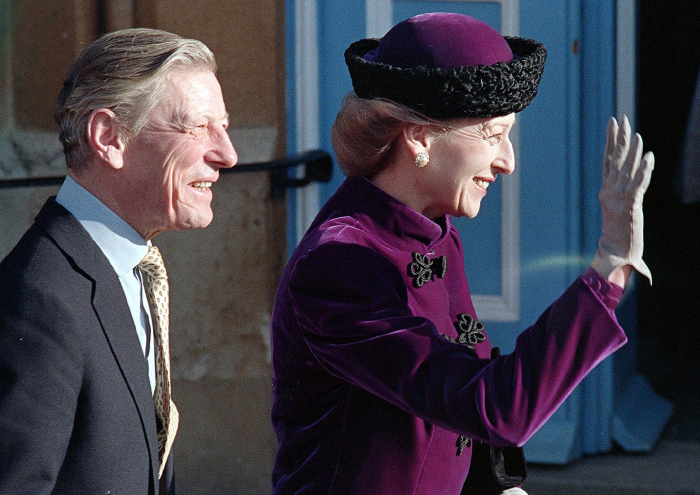 A man in a suit and a woman in purple coat and hat waving to someone off-camera