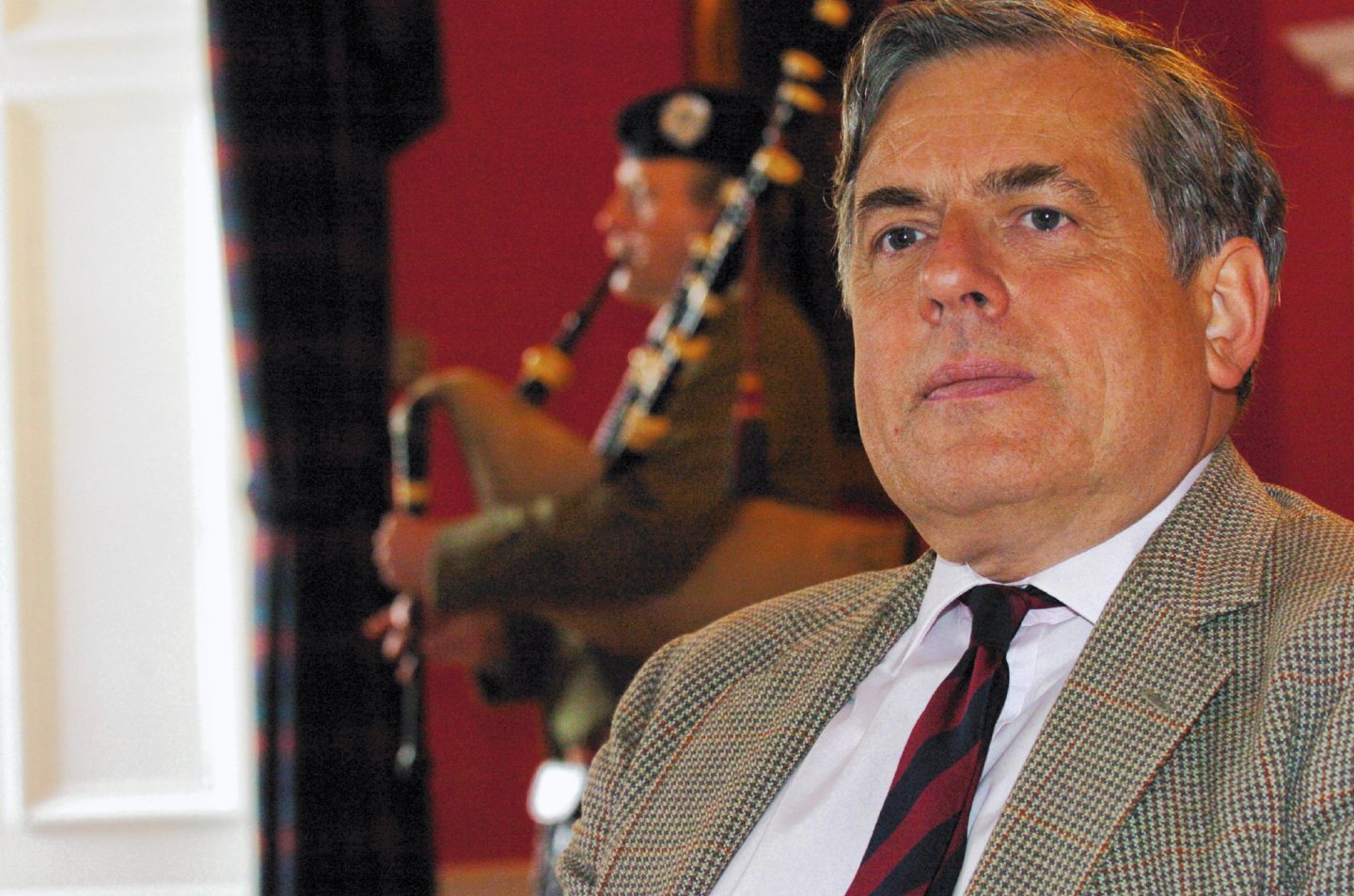 The head and shoulders of a man in shirt, tie and suit jacket with a piper playing in the background.