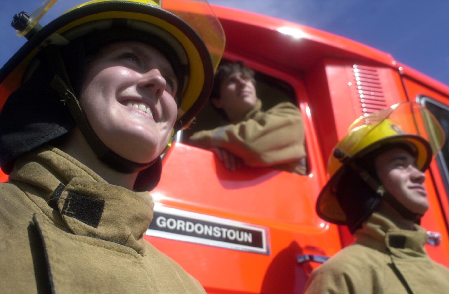 Three teenagers in fire uniform and helmets, one sitting in a fire engine