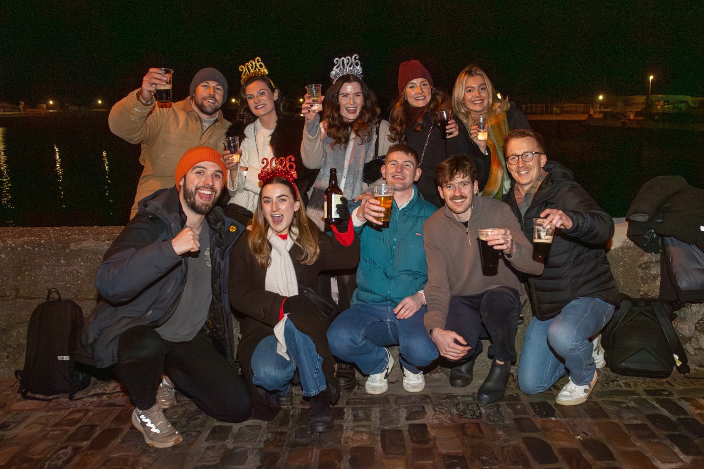 Group of friends celebrate Hogmanay by Stonehaven Harbour, raising drinks and wearing 2026 headbands during New Year celebrations in Stonehaven.