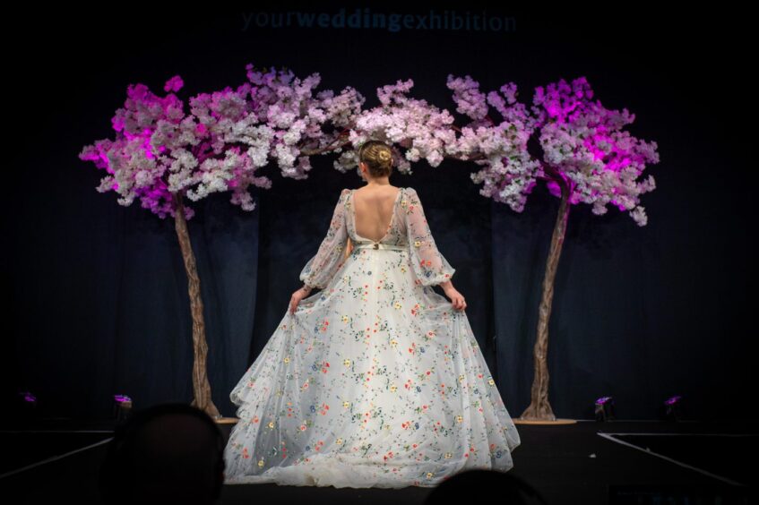 A model walking on the catwalk in a floral‑embroidered wedding gown beneath illuminated blossom trees during the 2026 Your Wedding Exhibition at P&J Live.