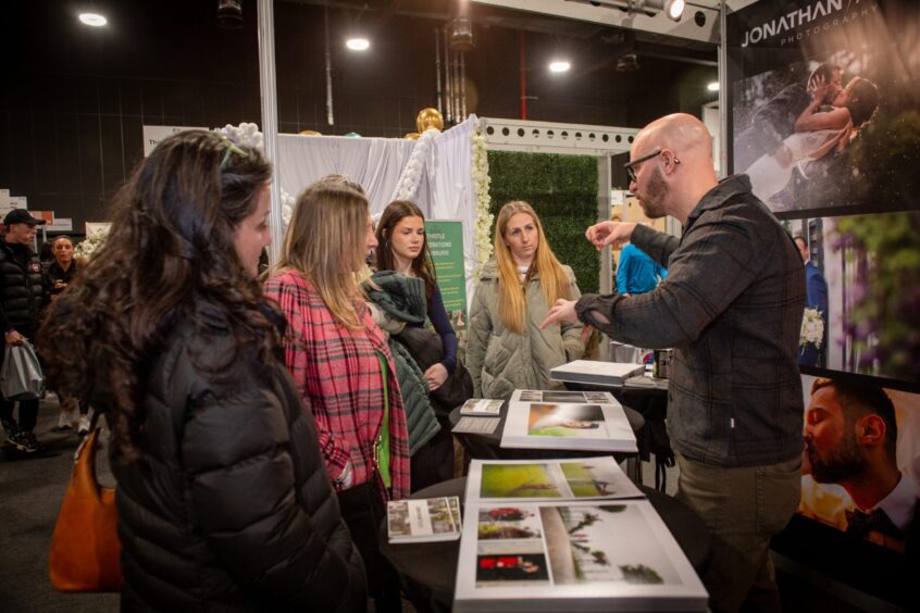 A wedding photographer showing sample photo albums to visitors at Your Wedding Exhibition 2026 inside P&J Live, Aberdeen.