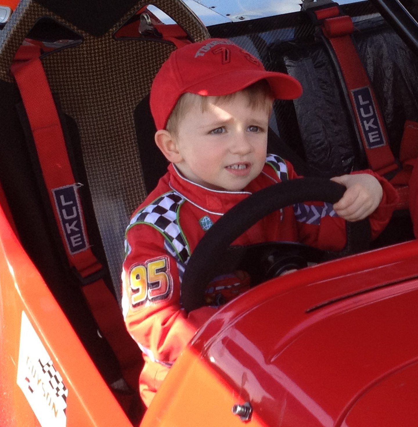 Euan as a little boy in a red cap and boilersuit sitting in the driver's seat of a red car.