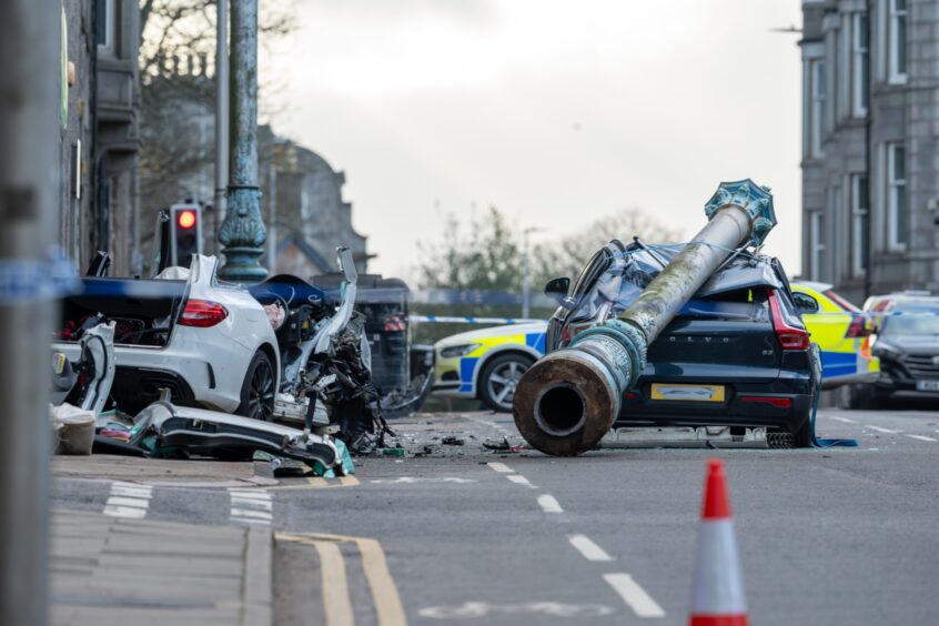 A severe car crash scene shows two heavily damaged cars on a street, with a fallen post across one vehicle and a police car behind.