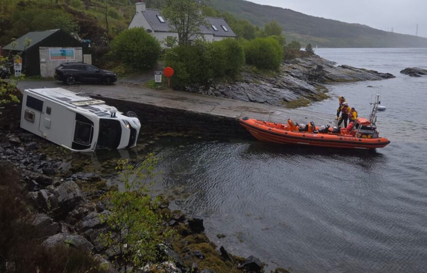 A white campervan lies overturned and partially submerged at a boat ramp. An orange RNLI rescue boat with crew is moored nearby, responding to the incident in Kyle Rhea narrows.