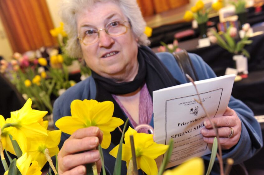 Mary McGregor at the Aboyne Spring Show, holding a yellow daffodil and a "Prize Schedule SPRING SHOW" booklet, surrounded by flower displays.
