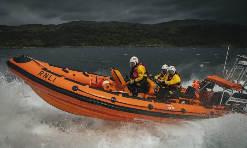 A bright orange RNLI rescue boat carrying three crew members in yellow gear speeds across choppy water with dark hills in the background.