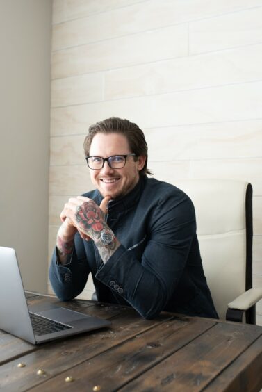 Man with glasses and tattooed hands sits behind laptop