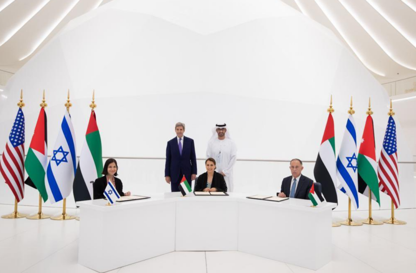 Three people sit behind desk, with flags, while two men stand at the back