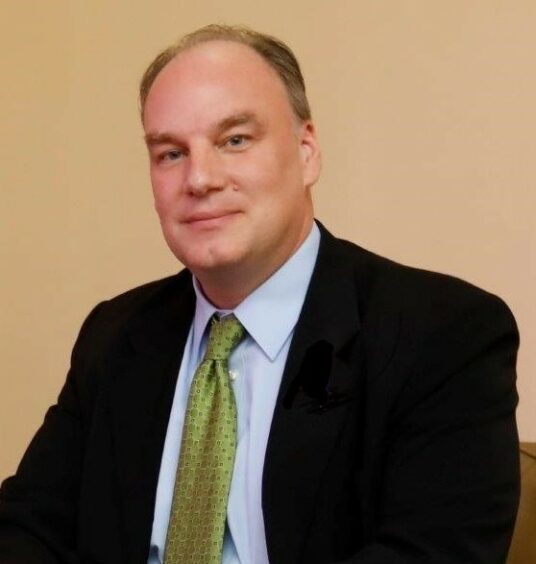 Headshot of man in black jacket, against pale brown background