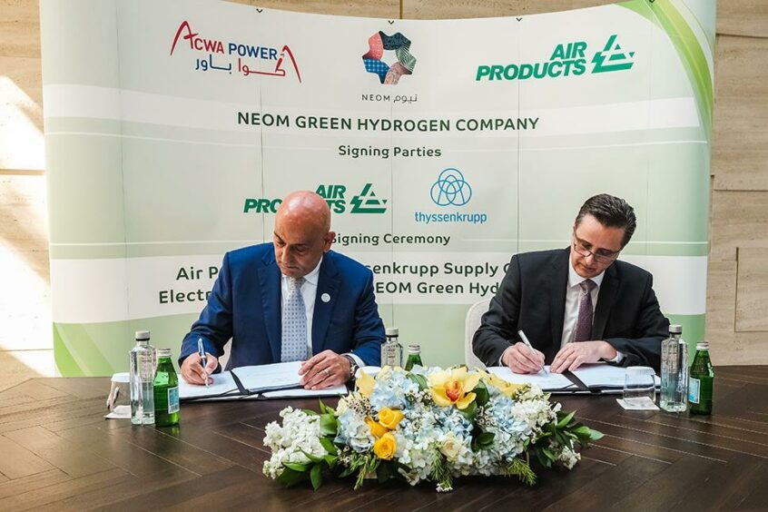 Two men sign documents on desk in front of logoed backdrop