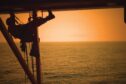 Rope worker dangling from scaffold above sea as sun sets