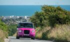 Pink car driving towards camera on a sunny day, wind turbines in the sea behind