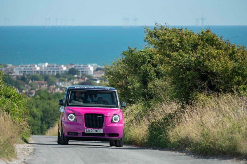 Pink car driving towards camera on a sunny day, wind turbines in the sea behind