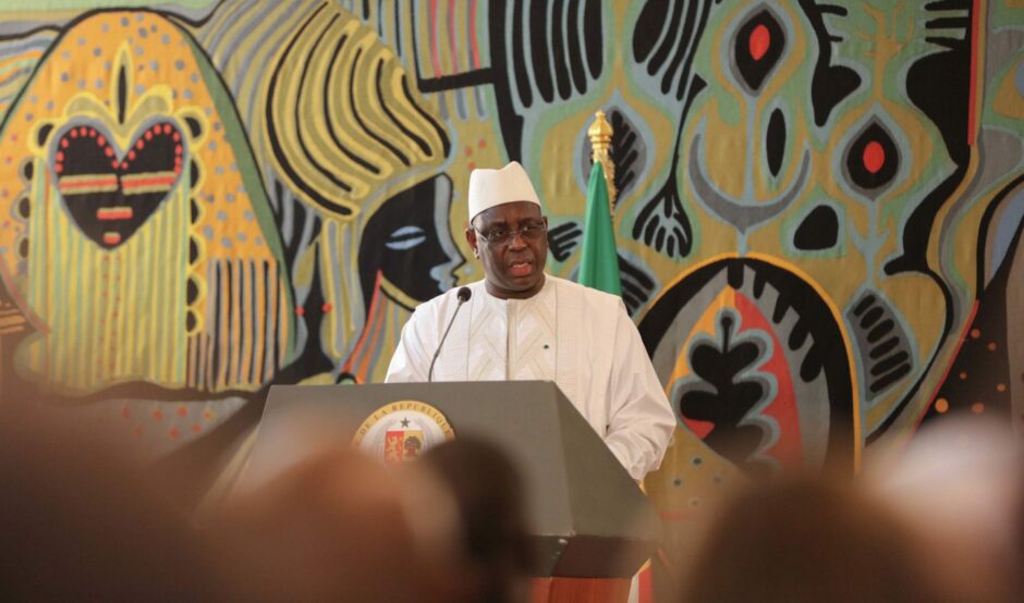 Man dressed in white stands in front of patterned wall, speaking into mic