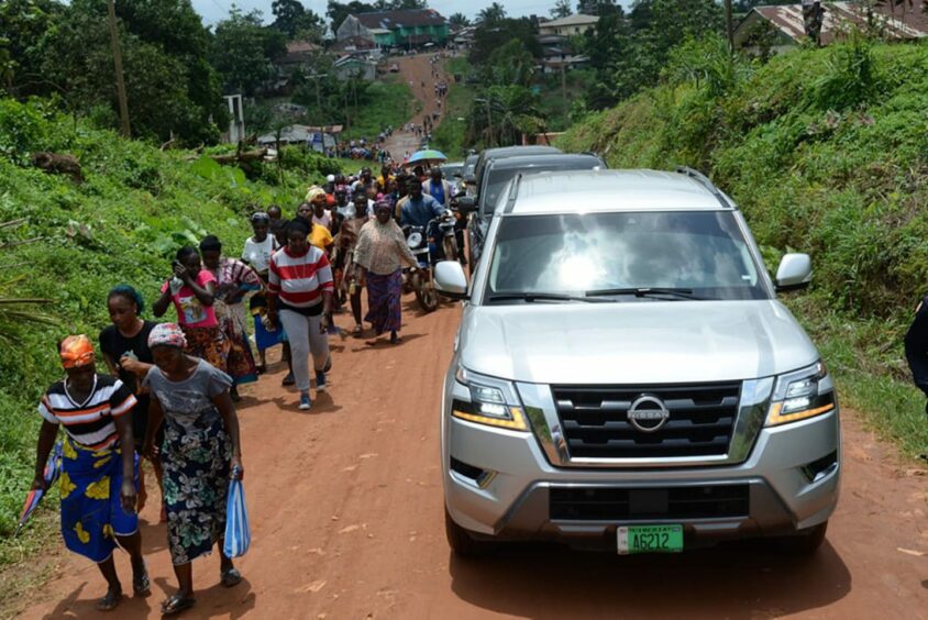 A shiny car drives past people walking on a dirt road