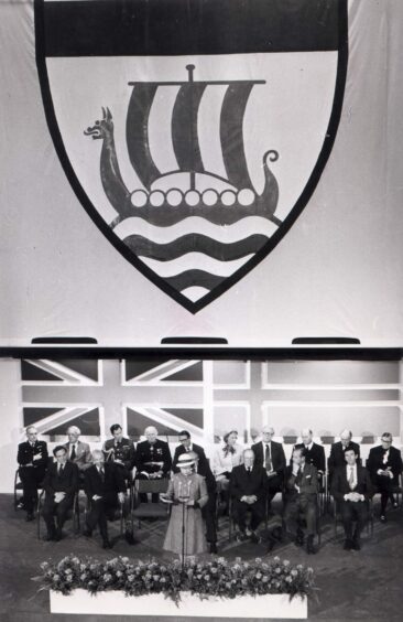 Members of the platform party listen to the Queen's speech at the Sullom Voe inauguration on a dias dominated by the terminal's Viking badge - May 1981