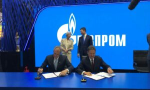 Two men sign documents in front of blue Gazprom sign
