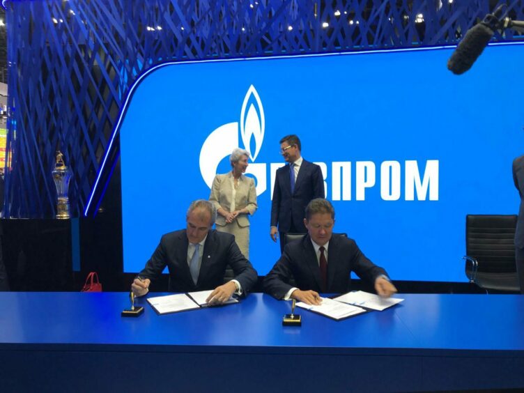 Two men sign documents in front of blue Gazprom sign