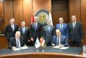 Group of men stand behind desk, two sign documents