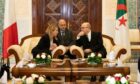 Two people talk, with another man behind, flanked by Italian and Algerian flags