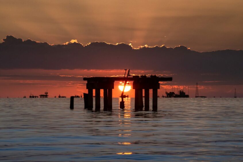 The sun sets behind a dilapidated oil rig in the sea.