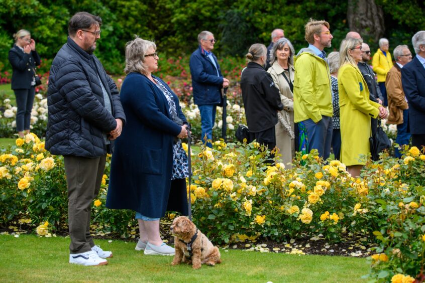 People and a small dog stand beside yellow rose bushes during the 2025 Piper Alpha remembrance service at Hazlehead Park in Aberdeen.