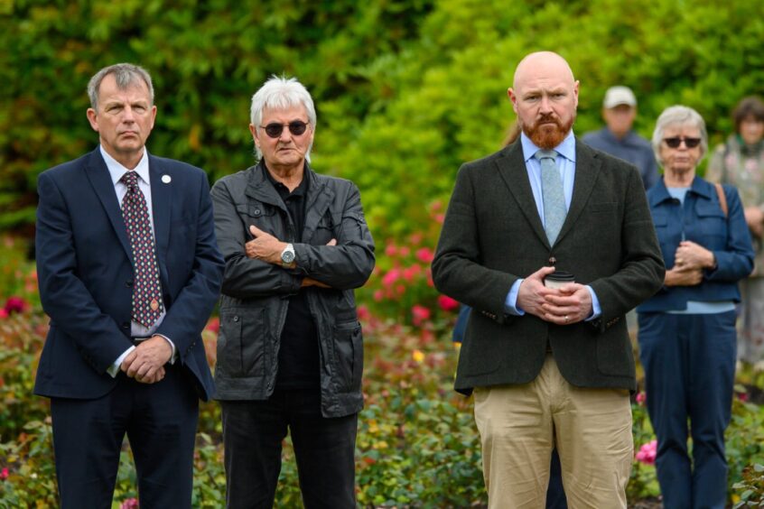 Three people stand in quiet reflection during the 2025 Piper Alpha memorial service at Hazlehead Park, Aberdeen, surrounded by others and blooming rose bushes.