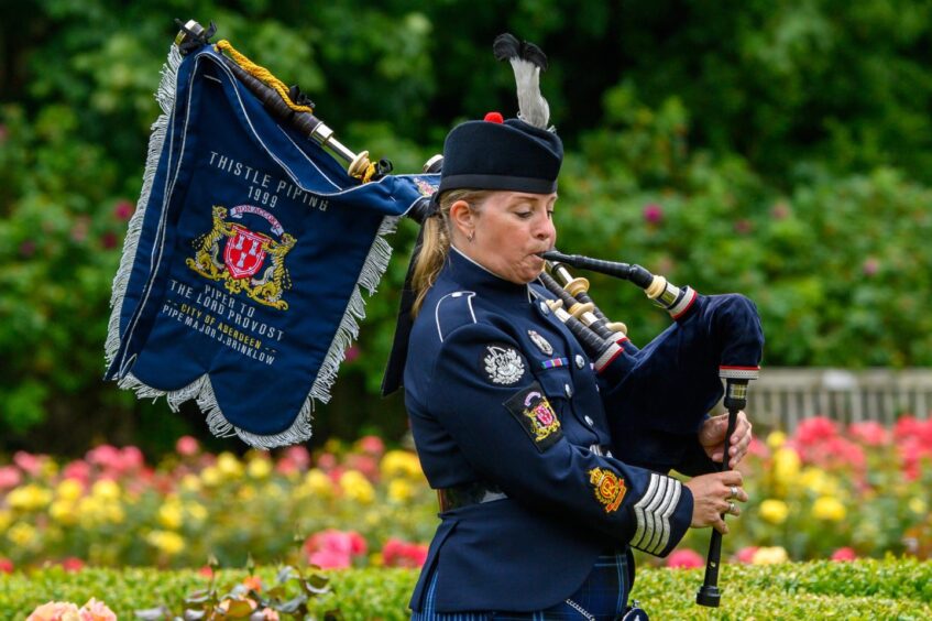 Piper Julie Brinklow plays the bagpipes in full uniform at the Piper Alpha memorial service in Hazlehead Park, Aberdeen, with floral gardens in the background.