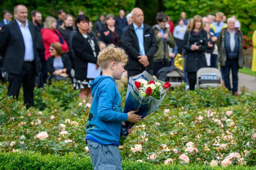Blond boy in blue hoodie holding a bouquet of red and white roses, surrounded by rose bushes and attendees at the Piper Alpha memorial service in Aberdeen.