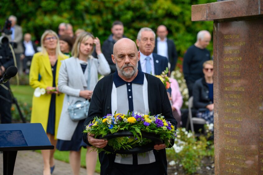 Man carrying a floral wreath approaches the Piper Alpha memorial, with others solemnly following behind during the commemorative event.