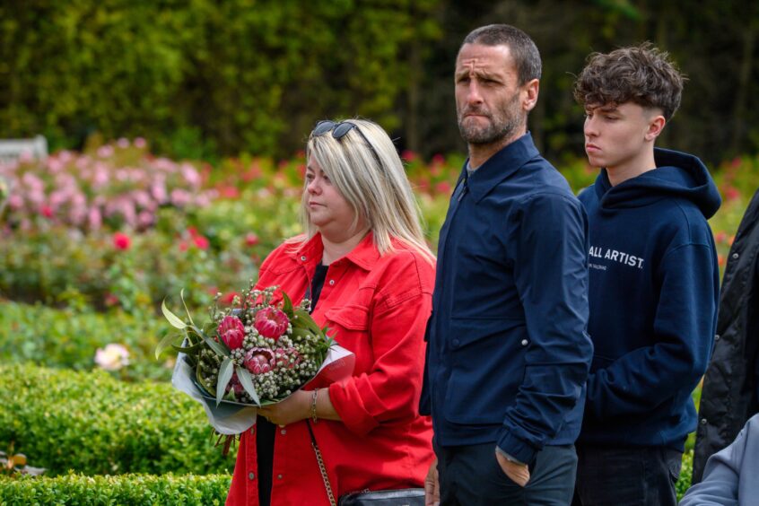 Three solemn individuals, including a woman holding a bouquet, stand in a rose garden during the Piper Alpha memorial service.