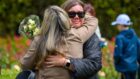 Two women hug during an emotional moment at the Piper Alpha memorial, one holding a bouquet of small white and pink flowers, with blurred crowd and rose bushes in the background.