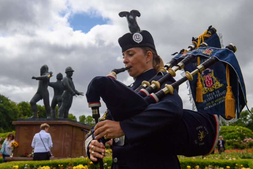 Piper Julie Brinklow plays the bagpipes in full uniform beside the Piper Alpha memorial statue in Hazlehead Park, with cloudy skies overhead.