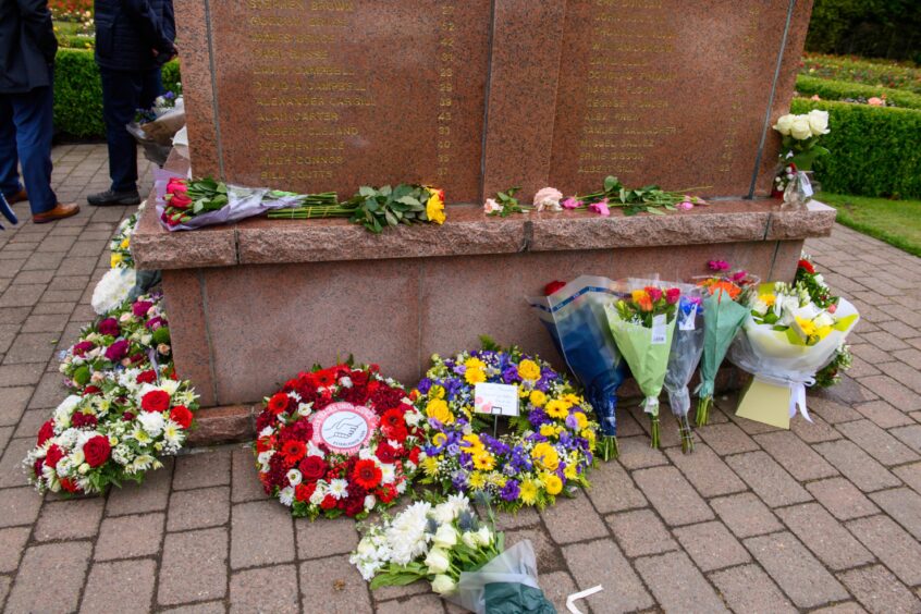 Bouquets and wreaths placed at the base of the Piper Alpha memorial in Hazlehead Park, with engraved names of victims visible on the stone monument.