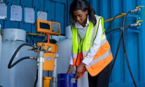 A young Black woman, dressed in professional attire, is filling liquefied petroleum gas into a client's tank at a filling station