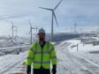 A man stands in the foreground with large onshore wind turbines dotted across a snowy mountainous landscape behind him, presumably in Scotland.
