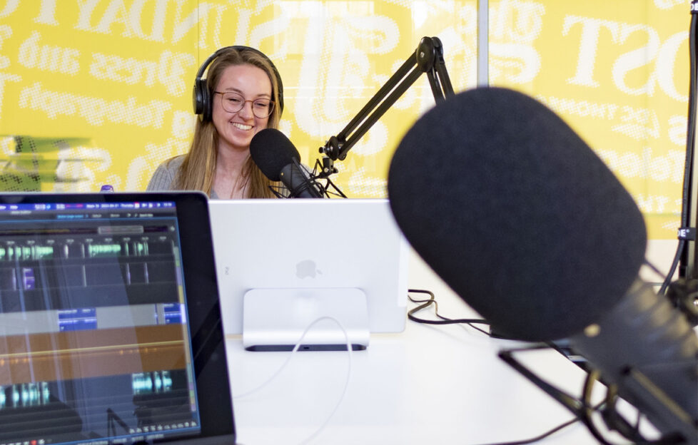 Abbie at podcast studio with editing screen and mic in foreground