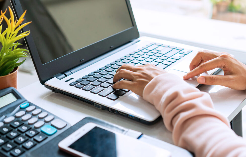 Woman's hands working at a laptop, next to her phone, calculator and small plant