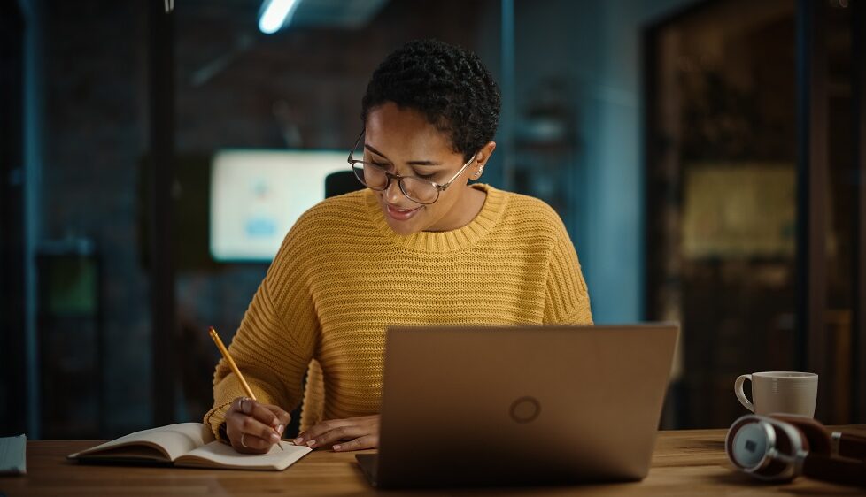 Woman writing in notepad with laptop at desk