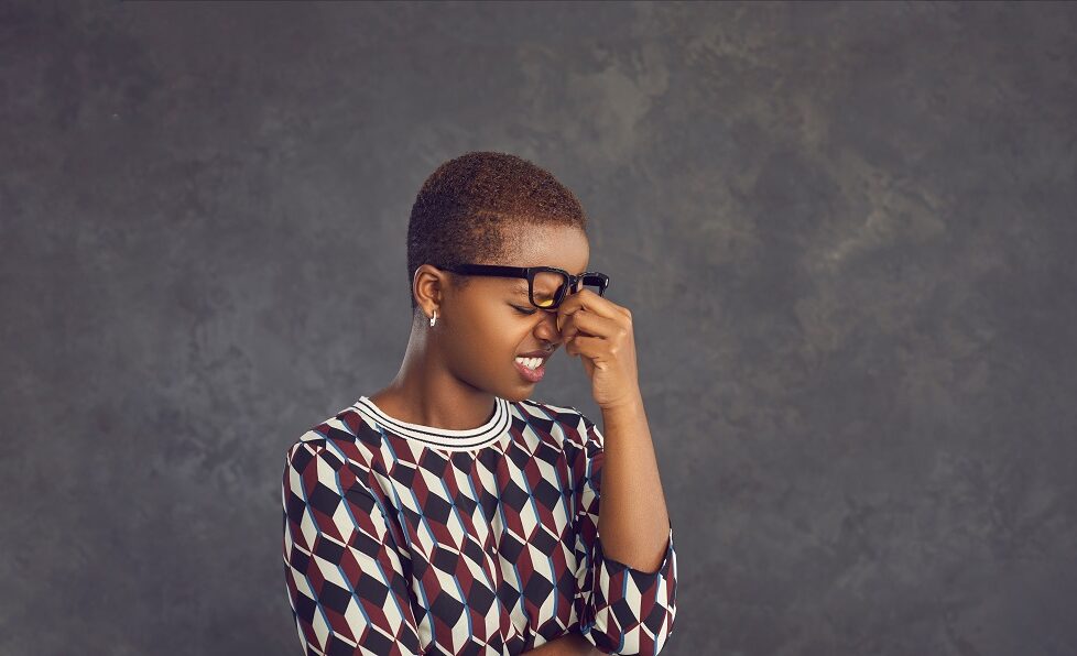 Black woman with close shaven hair pinching her nose looking stressed below her glasses, baring her teeth
