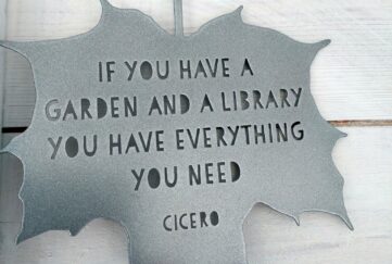 A metal silver hanging ornament in the shape of a leaf reading 'If you have a garden and a library you have everything you need', Cicero quote