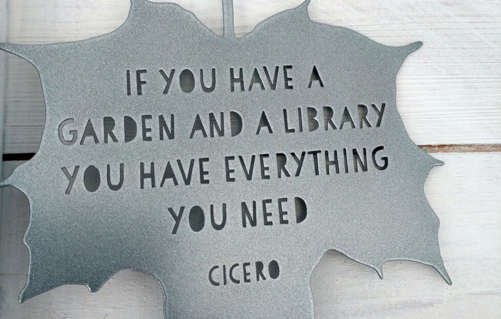 A metal silver hanging ornament in the shape of a leaf reading 'If you have a garden and a library you have everything you need', Cicero quote