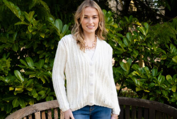 Model wearing a knitted cream cardigan with hooped gold necklace and blue jeans on foliage background