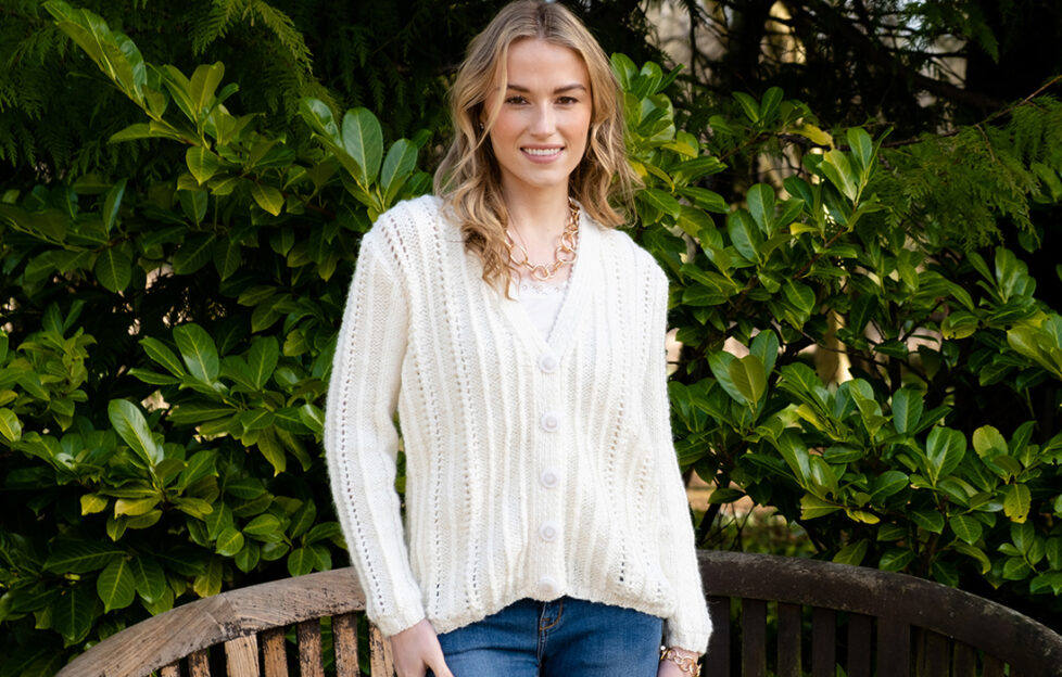 Model wearing a knitted cream cardigan with hooped gold necklace and blue jeans on foliage background