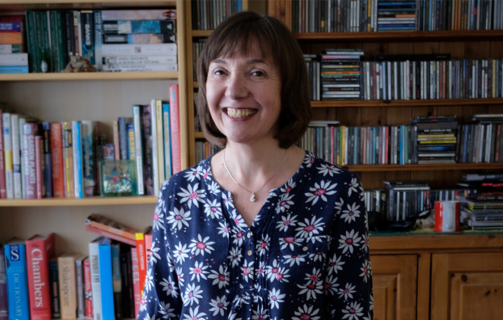 Headshot of Glenda Young sitting in front of a wall of bookshelves, smiling to the camera