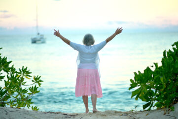 Elderly woman standing on beach facing the sea with arms raised towards the sunset