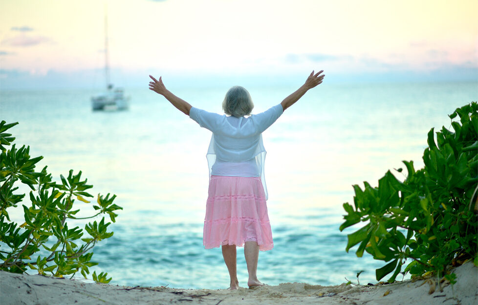 Elderly woman standing on beach facing the sea with arms raised towards the sunset
